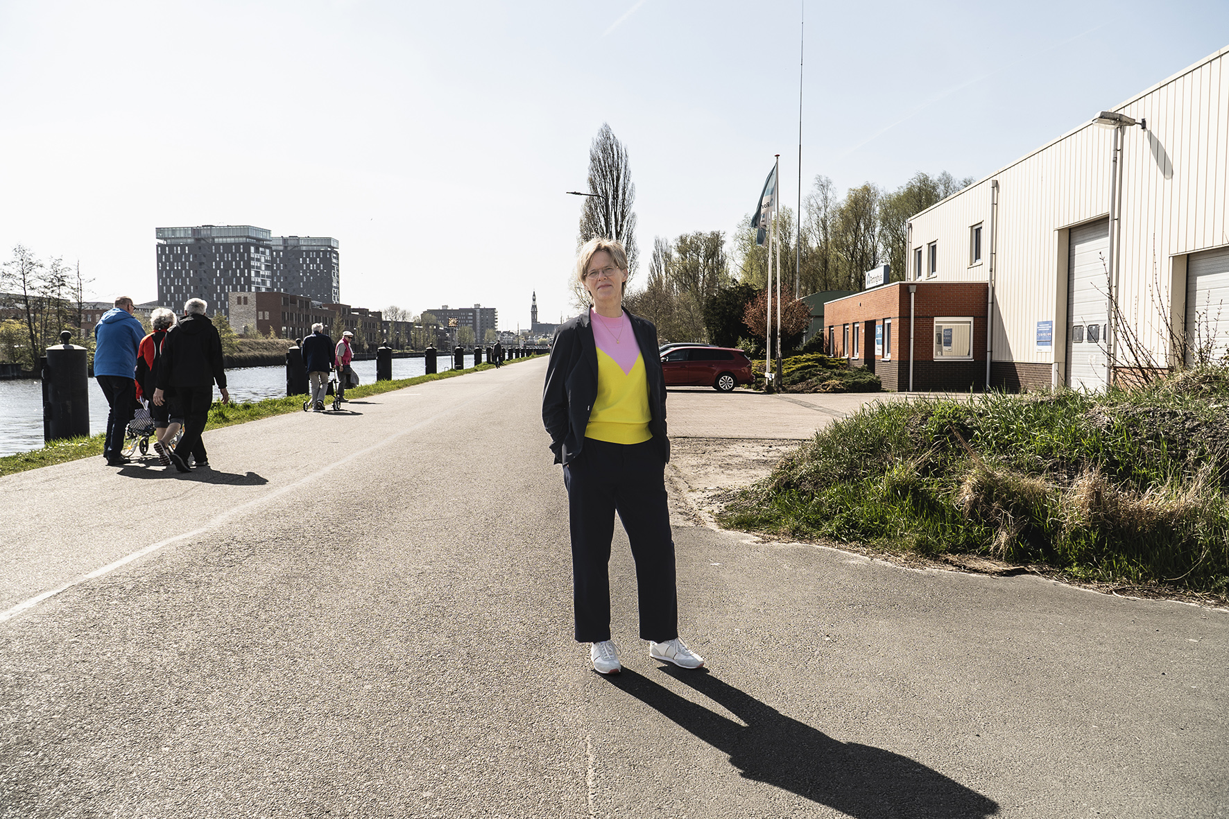Foto van vrouw met roze geel shirt, zwart pak en witte schoenen naast het water ( damsterdiep )